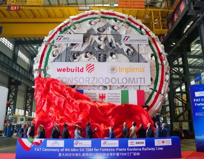 Wide ceremony view of the ribbon-draped single-shield cutterhead with attendees lined up in front of the machine inside the factory hall