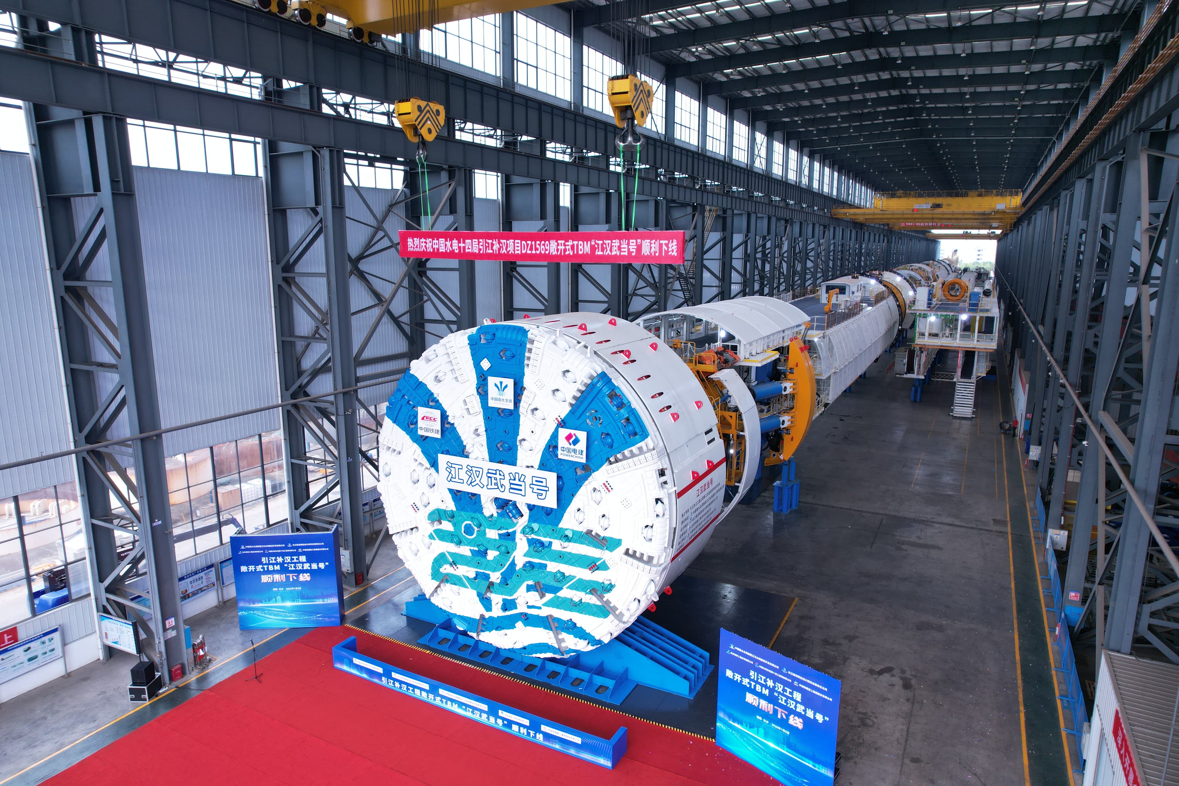Jianghan Wudang 12 metre-class TBM at roll-off with the "江汉武当号" banner across the cutterhead, red ceremonial mat in the foreground and the machine body extending down the factory hall